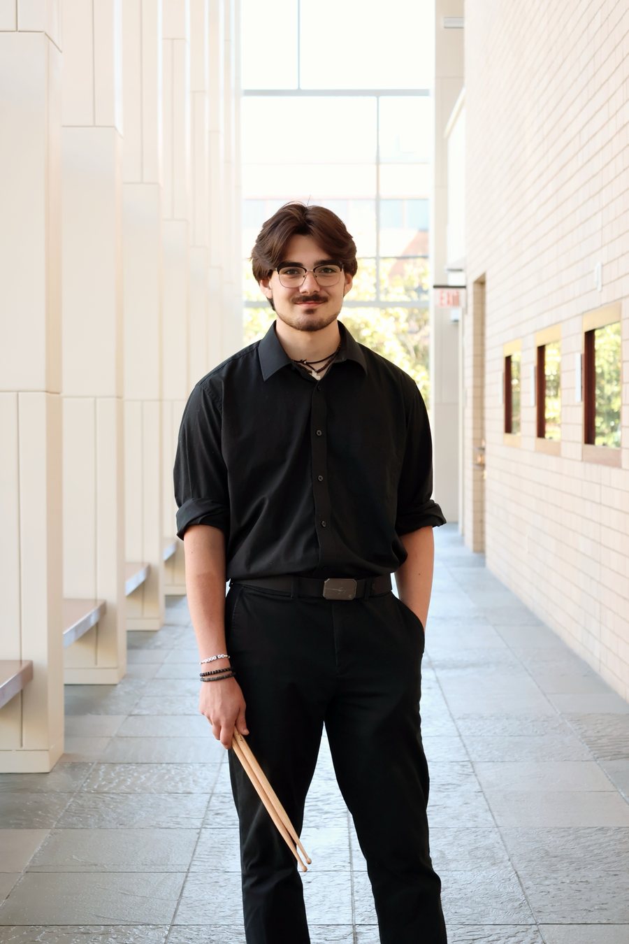 Makoa Lewis in a black shirt, holding drumsticks, standing in a sunlit hallway